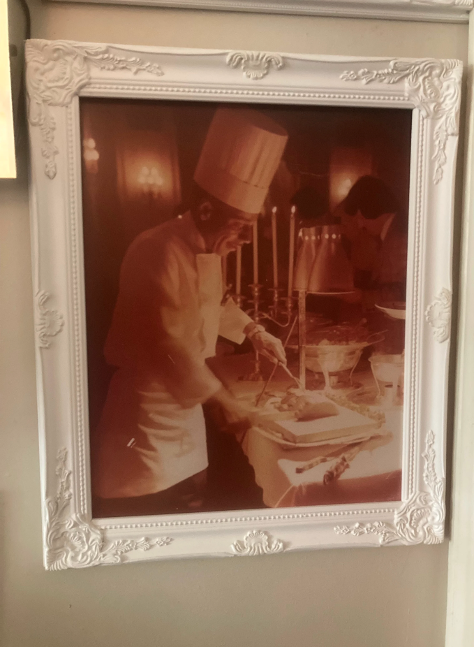 Framed sepia-toned photograph of a chef in a tall hat carving food at a candlelit table.
