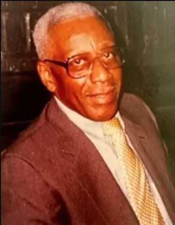 Middle-aged man with gray hair and glasses wearing a brown suit and yellow tie.