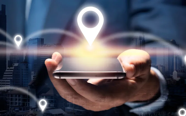 Hand holding smartphone with glowing GPS location icons overlaid on cityscape background.