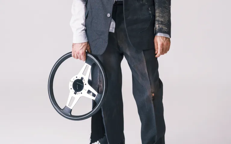 Man wearing a dirty, torn suit holding a detached car steering wheel against a plain background.