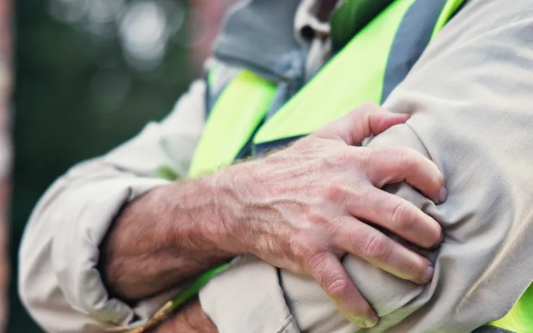 Close-up of a person in a beige shirt and neon safety vest clutching their left upper arm.