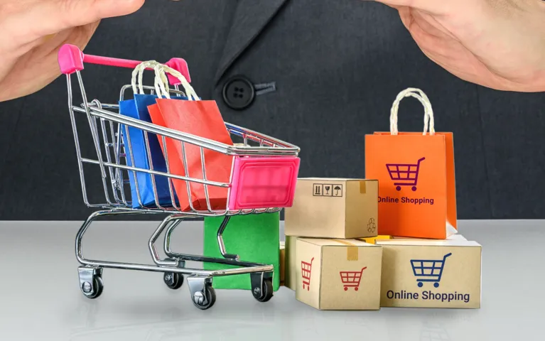 Person in dark suit gestures over miniature shopping cart with colorful bags and cardboard boxes labeled "Online Shopping.