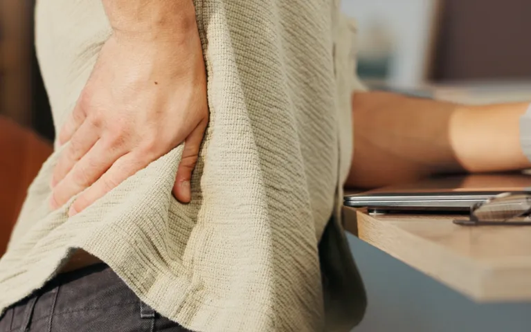 Person wearing a beige sweater holding their lower back near a wooden desk.