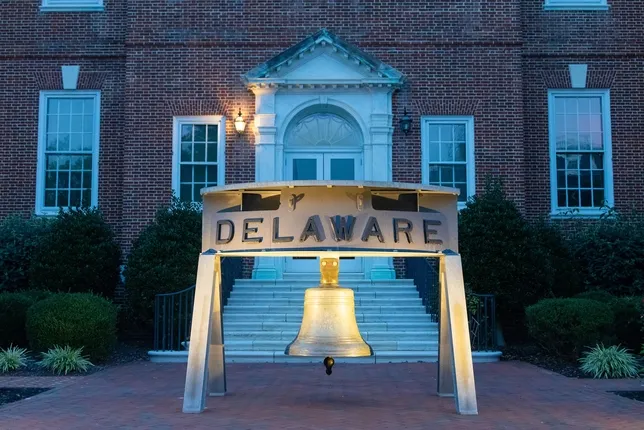 Illuminated bronze bell with "DELAWARE" inscription in front of red brick building with white entrance.