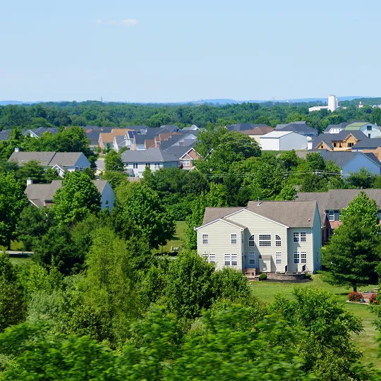 A suburban neighborhood with multiple houses surrounded by dense green trees under a clear blue sky.