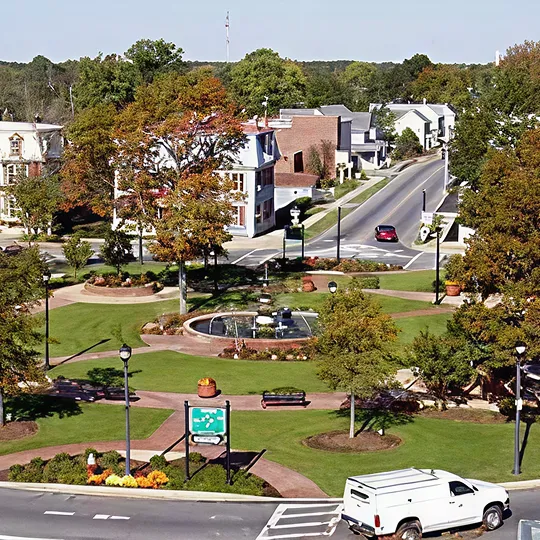 A small town park with a central fountain, autumn trees, paved walkways, benches, and surrounding streets with vehicles.