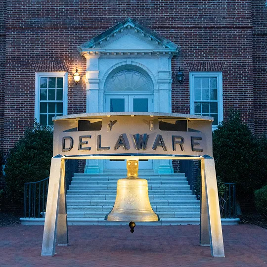 Illuminated metallic Delaware bell sculpture in front of brick building with white staircase.