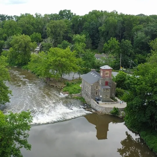 Aerial view of a stone building beside a flowing river surrounded by dense green trees.